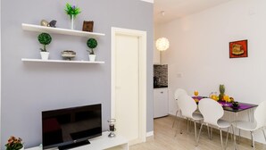 A modern living room with a flat-screen TV, white shelves with decorative items, a dining area with a table and chairs, and a kitchen in the background.