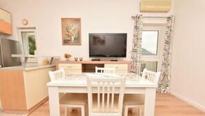 A dining area with a white table and chairs, a flat-screen TV on a wooden cabinet, and a window with floral curtains. - Green Hills Apartment - One Bedroom Apartment With Balcony and Sea View (Dubrovnik)