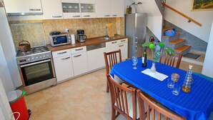 A modern kitchen with white cabinets, a gas stove, a microwave, and a coffee maker. There is a dining table with a blue tablecloth and chairs. A staircase leads to a lofted area. - Apartment Mambo , Mljet - Duplex Apartment With Terrace and Sea View (Mljet)