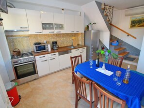 A modern kitchen with white cabinets, a gas stove, a microwave, and a coffee maker. There is a dining table with a blue tablecloth and chairs. A staircase leads to a lofted area. - Apartment Mambo , Mljet - Duplex Apartment With Terrace and Sea View (Mljet)