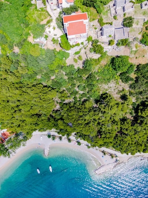 An aerial view of a coastal area with a beach, a small boat, and a building with a red roof. - Villa Kosa - Room With Balcony and sea View (Brela)