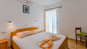 A bedroom with a wooden bed, a nightstand, a chair, a window with curtains, and an air conditioning unit. - Villa Kosa - Room With Balcony and sea View (Brela)