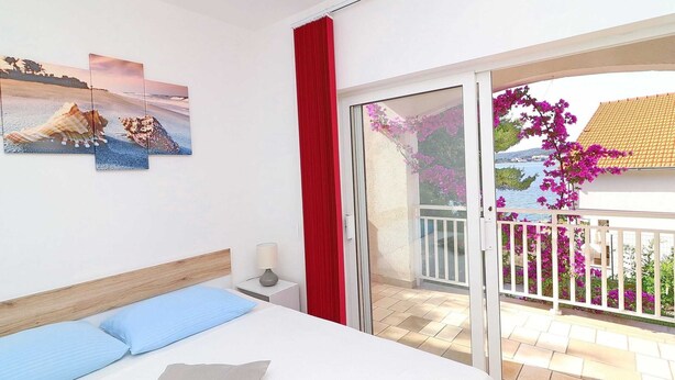 Et soveværelse med en seng, et natbord, en lampe og en skydedør, der fører ud til en balkon med blomster og havudsigt.