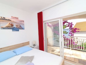 A bedroom with a bed, a nightstand, a lamp, and a sliding glass door leading to a balcony with flowers and a view of the sea. - Apartments Marko Peljesac - Standard One Bedroom Apartment With Terrace (Orebic)