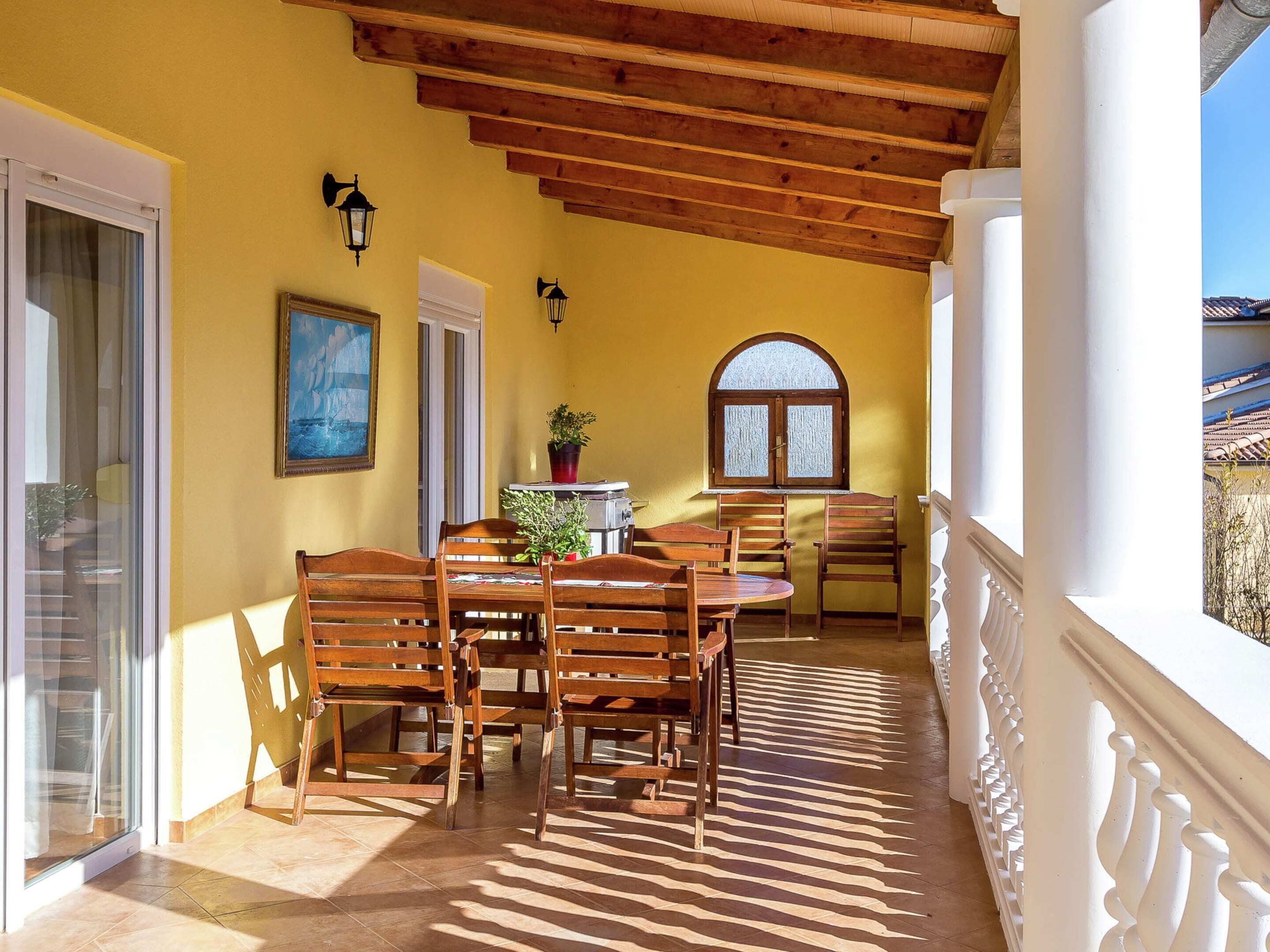 Une terrasse ensoleillée, meublée de mobilier en bois, agrémentée d’une plante en pot et offrant une vue sur des bâtiments traditionnels.