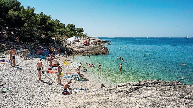Una playa con agua cristalina, costa rocosa y gente nadando y tomando el sol.