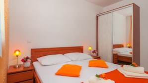 Una cama bien hecha con almohadas blancas y naranjas, un cabecero de madera y un espejo grande. Hay dos mesitas de noche con lámparas y un jarrón con flores.