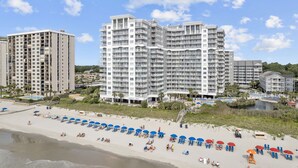 A beachfront hotel with multiple high-rise buildings, a sandy beach with umbrellas, and a clear blue sky. - Oceanfront Winter Escape- Ask About Monthly Discounts – Heated Pools/Spas/Views (Myrtle Beach)