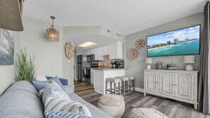 A modern living room with a sofa, ottomans, a flat-screen TV, and a kitchen in the background. - Oceanfront Winter Escape- Ask About Monthly Discounts – Heated Pools/Spas/Views (Myrtle Beach)
