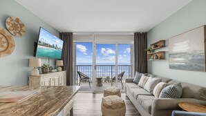 A living room with a TV, sofa, ottoman, and a balcony with a view of the sea. - Oceanfront Winter Escape- Ask About Monthly Discounts – Heated Pools/Spas/Views (Myrtle Beach)