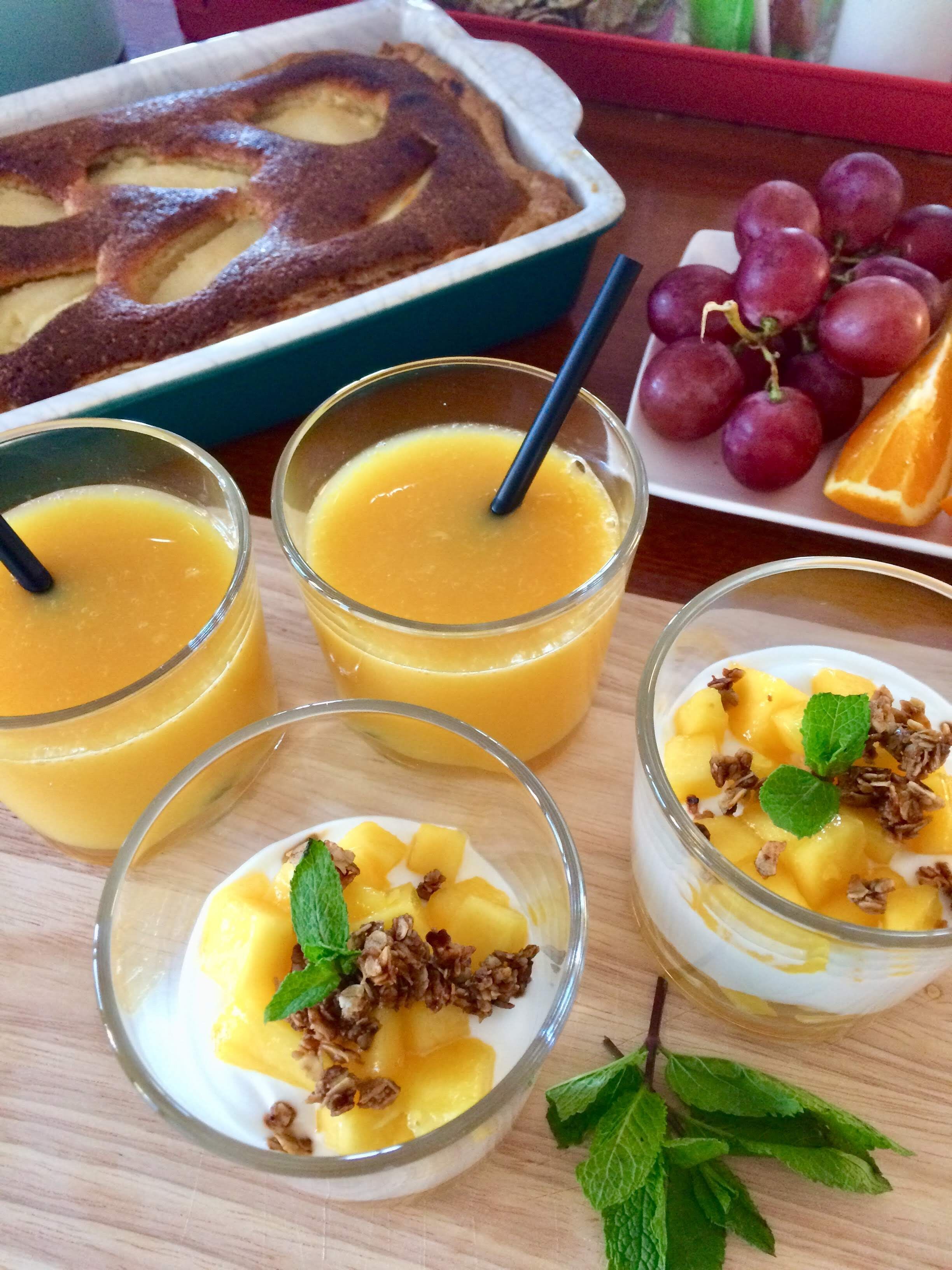 Three glasses of mango juice with straws, a plate of grapes and an orange slice, and a dessert with mango chunks and granola.
