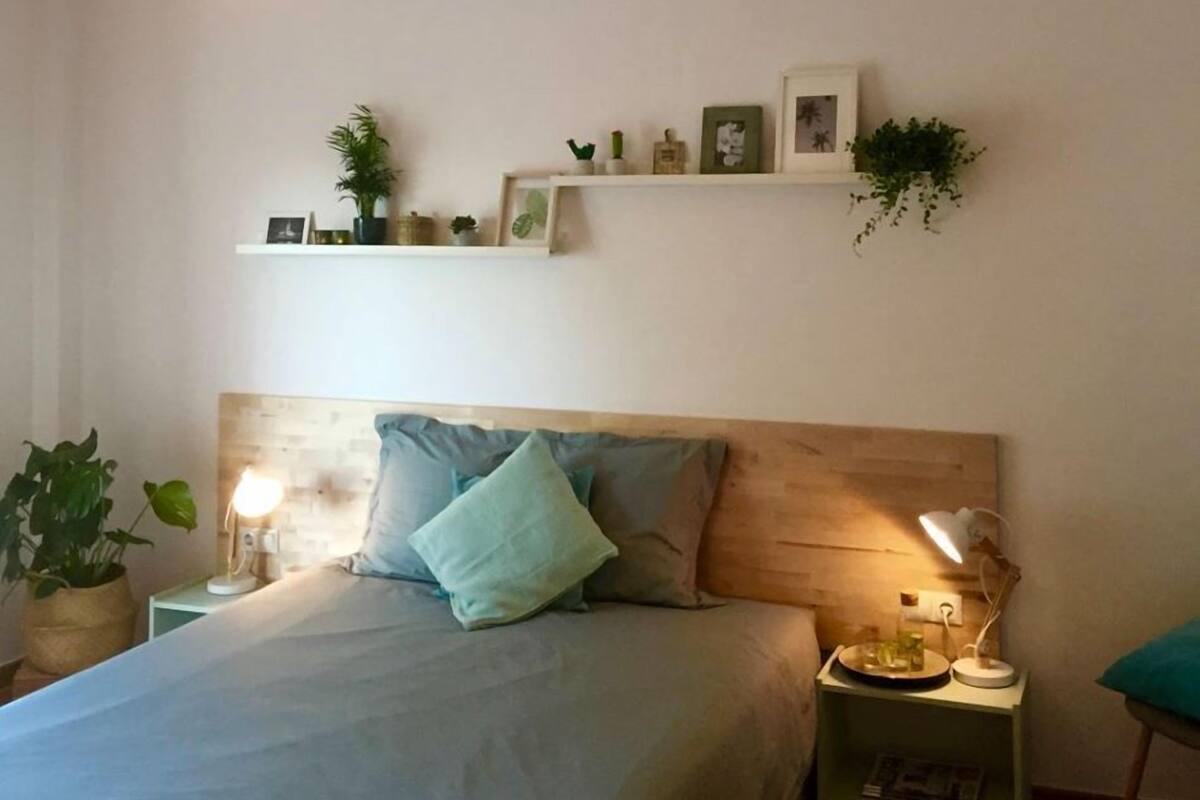 A bedroom with a bed, bedside tables, a hanging light fixture, and wall-mounted shelves with decorative items.