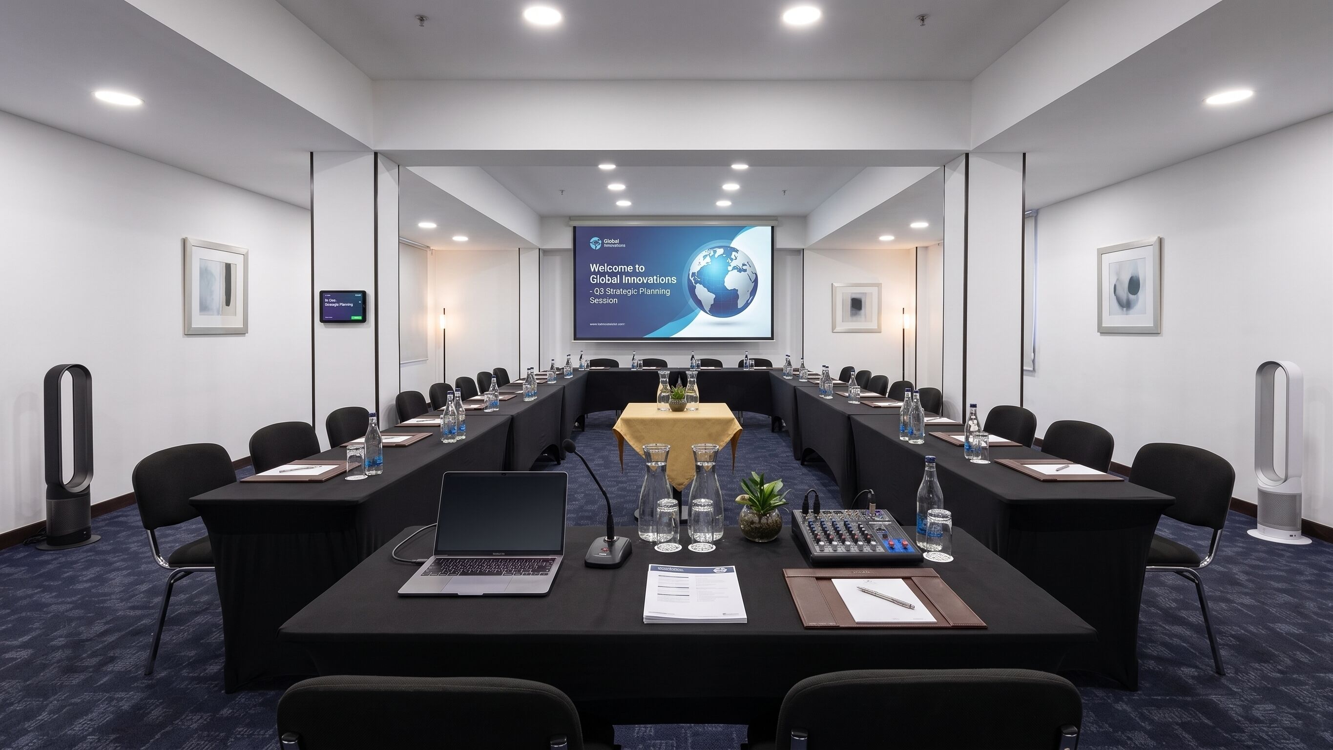 Un salón de conferencias con una larga mesa, sillas, computadoras portátiles, botellas de agua y una pantalla de presentación.