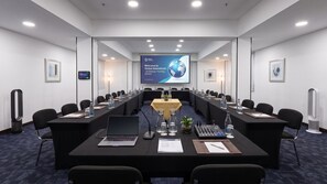 Un salón de conferencias con una larga mesa, sillas, computadoras portátiles, botellas de agua y una pantalla de presentación.