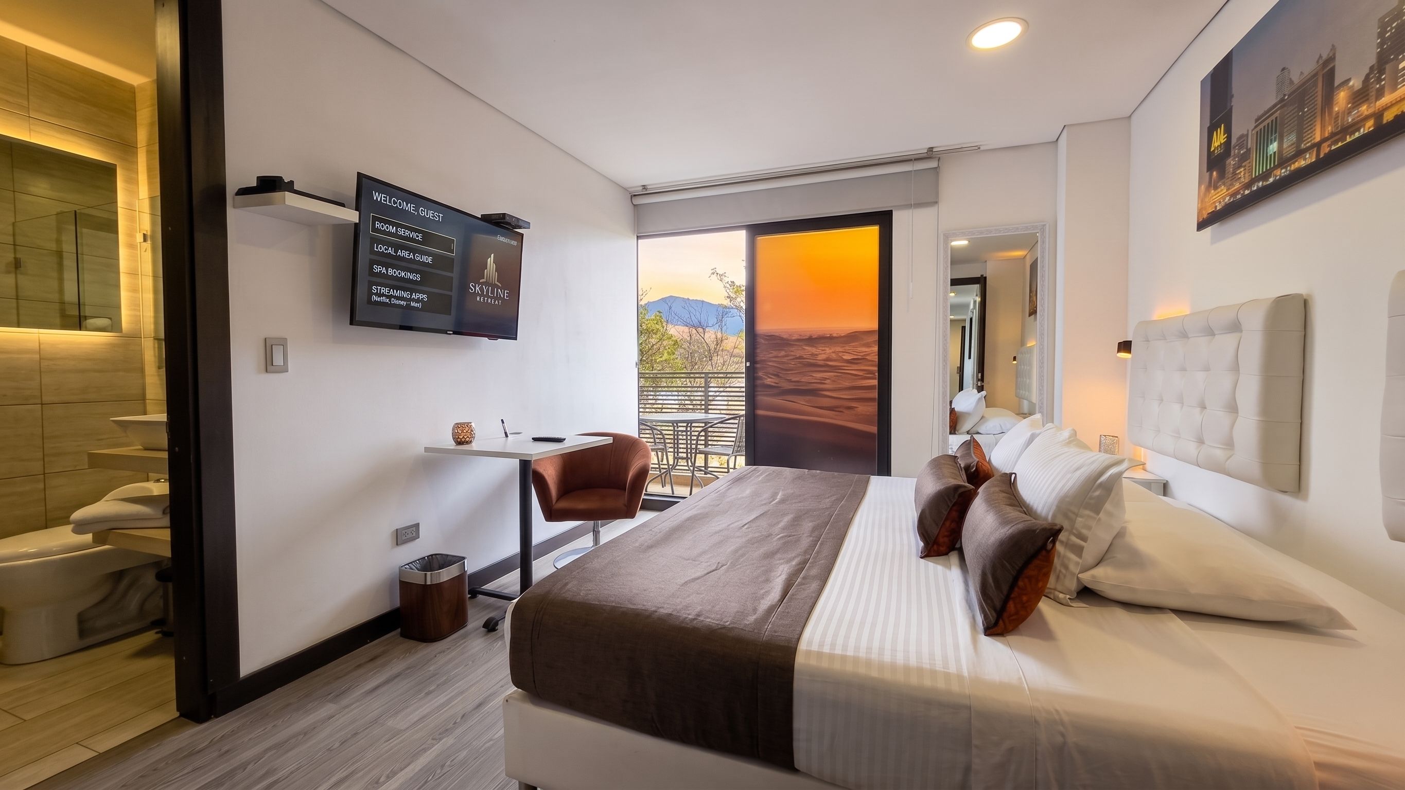 A modern hotel room with a bed, a desk, a chair, a TV displaying a welcome message, and a balcony with a desert view.