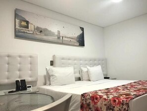Una habitación de hotel con una cama grande, una mesa redonda de vidrio, un cabecero blanco y un cuadro de un paisaje urbano en la pared.