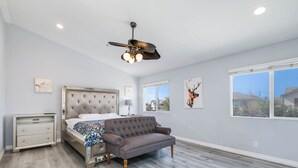 Une chambre à coucher avec un lit, un canapé gris, une table de chevet, un ventilateur de plafond et des œuvres d’art accrochées aux murs.