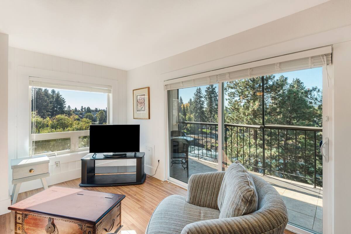 A modern living room with a flat-screen TV, a sofa, a wooden coffee table, and a view of a balcony with trees.