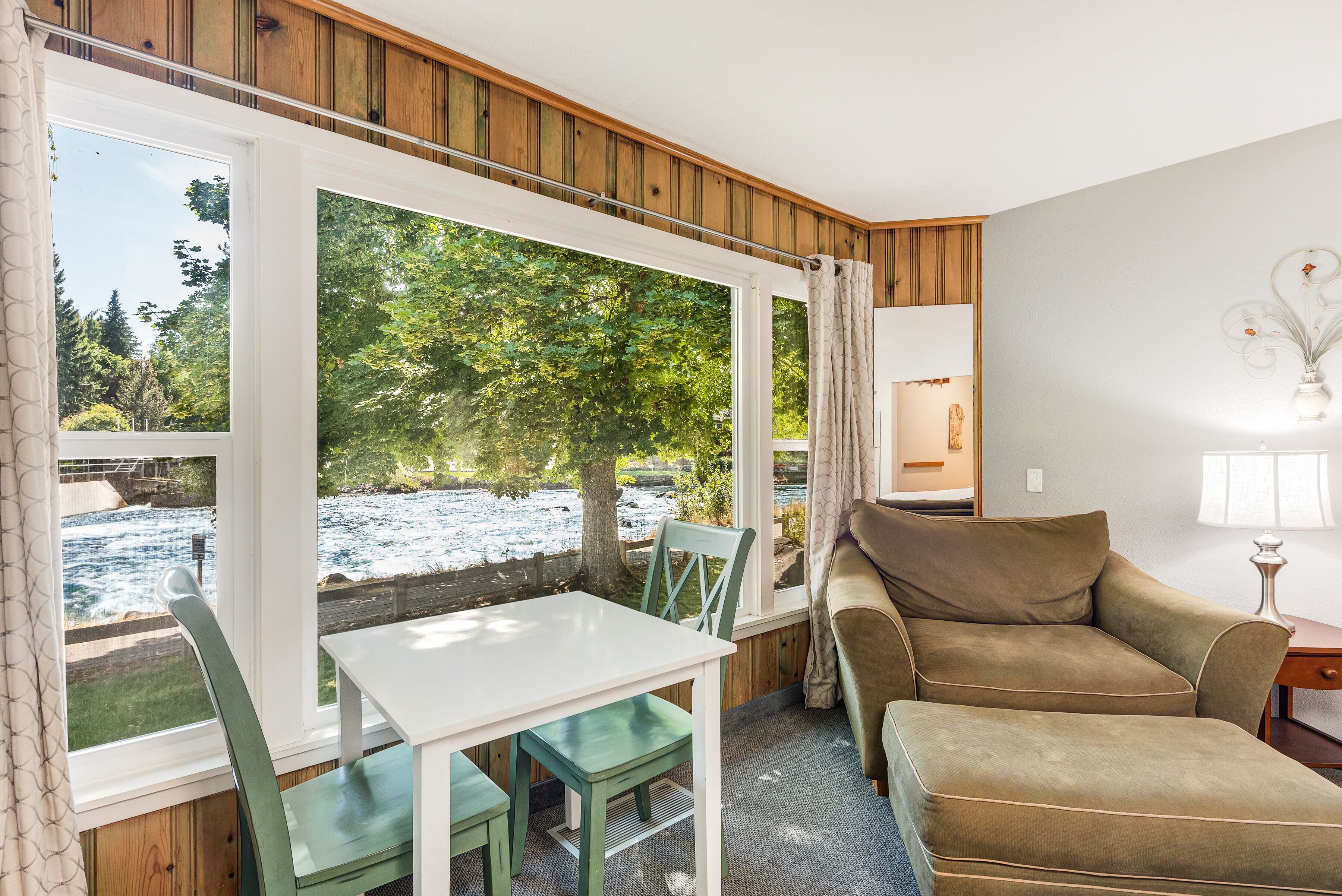 Une pièce avec un plafond en bois, une table blanche avec des chaises, un canapé marron et une grande fenêtre donnant sur une rivière et des arbres.