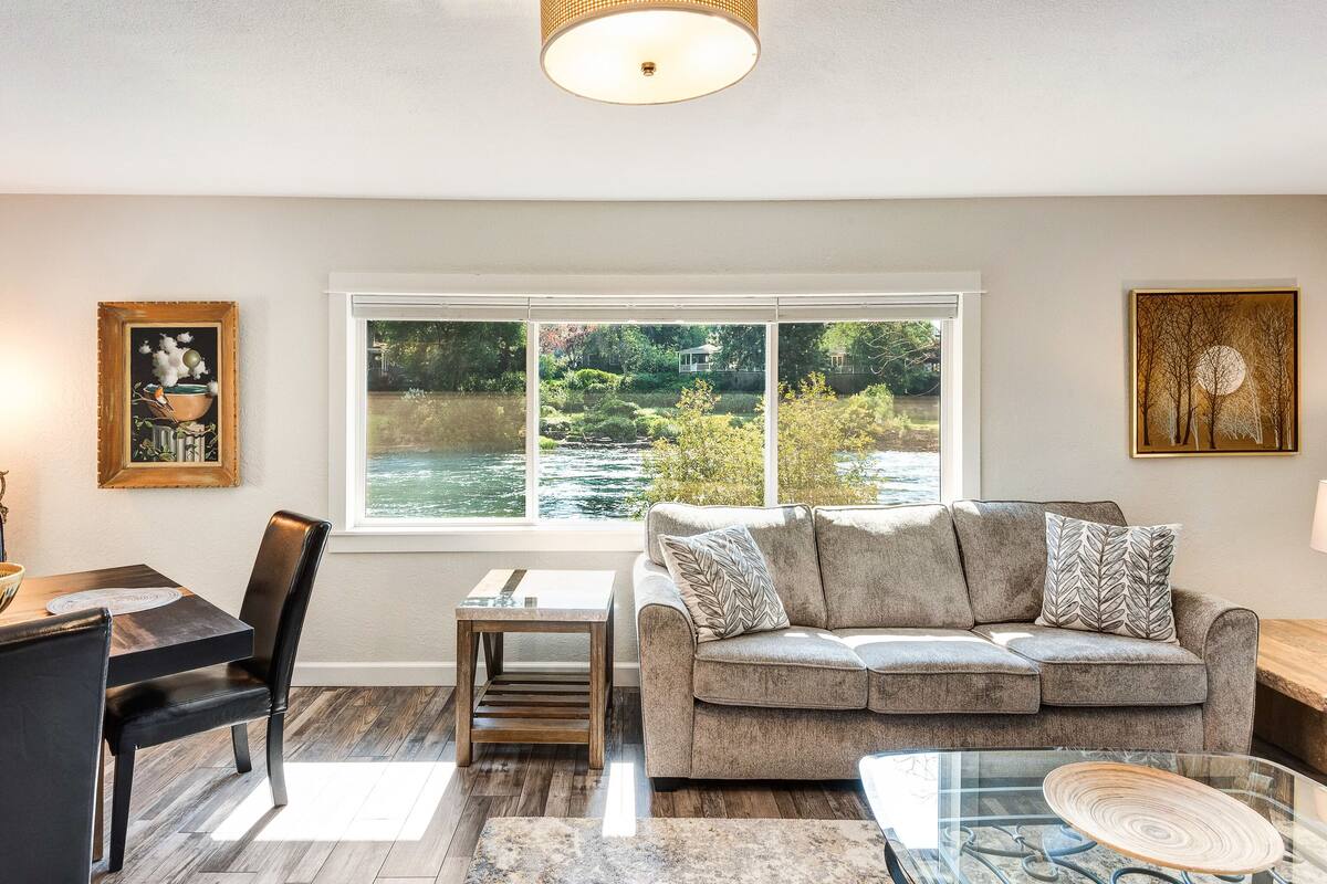 A living room with a view of a river, a dining area with a table and chairs, and a sofa with decorative pillows.