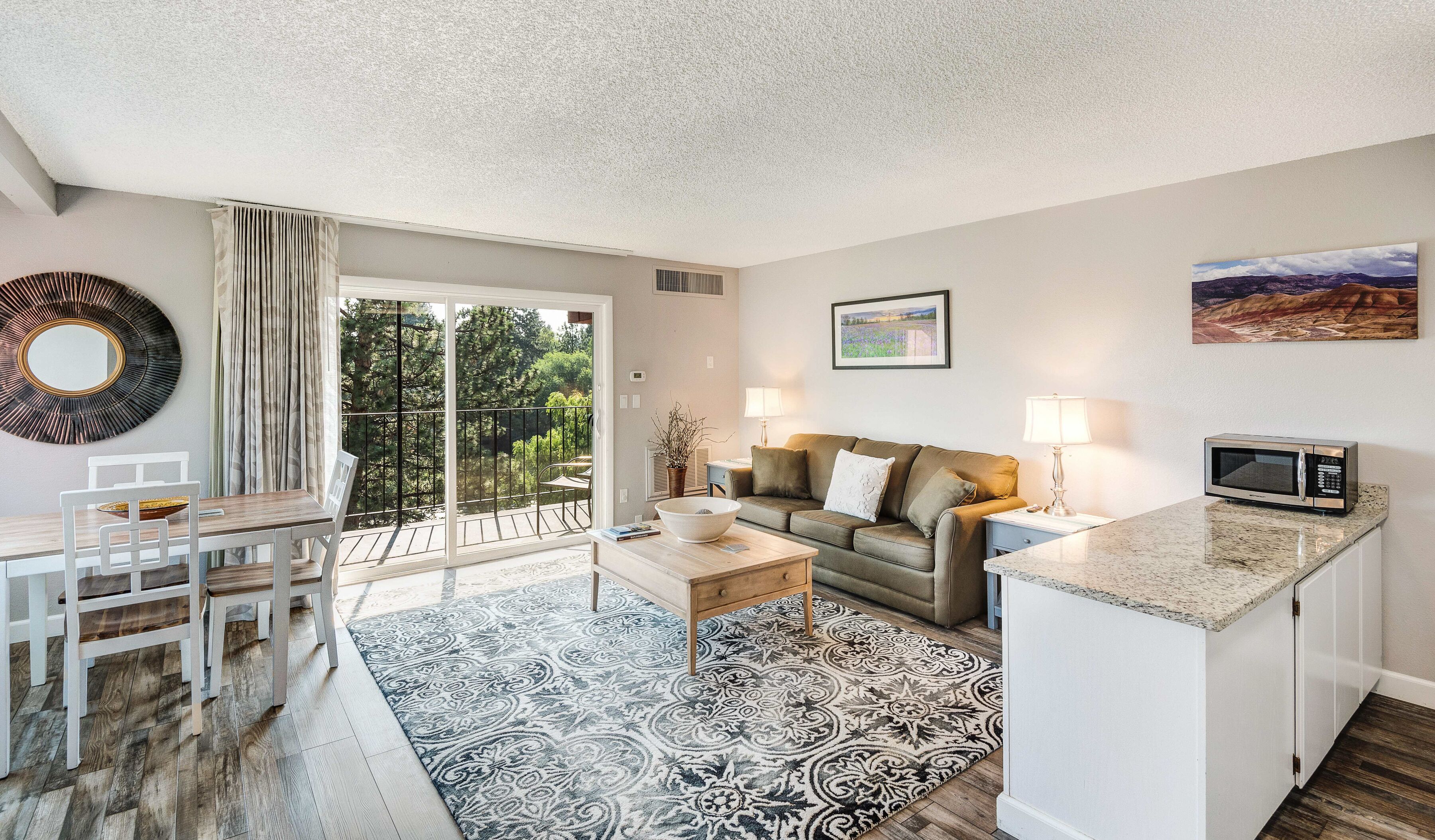 A living room with a sofa, a dining table with chairs, a coffee table, a kitchen area with a microwave, and a view of the outdoors through a sliding door.