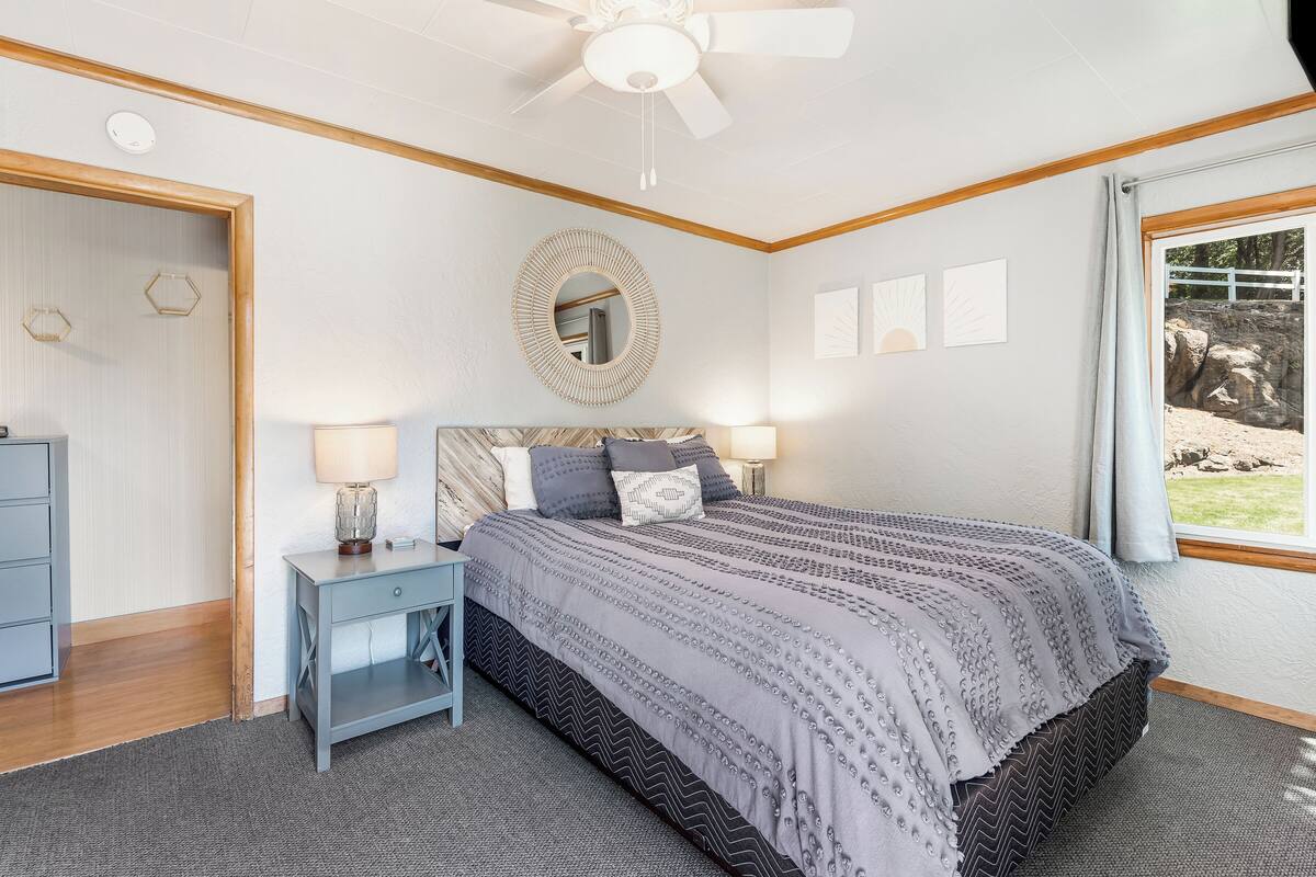 A bedroom with a bed, nightstands, a ceiling fan, a mirror, and a window with a view of rocks.