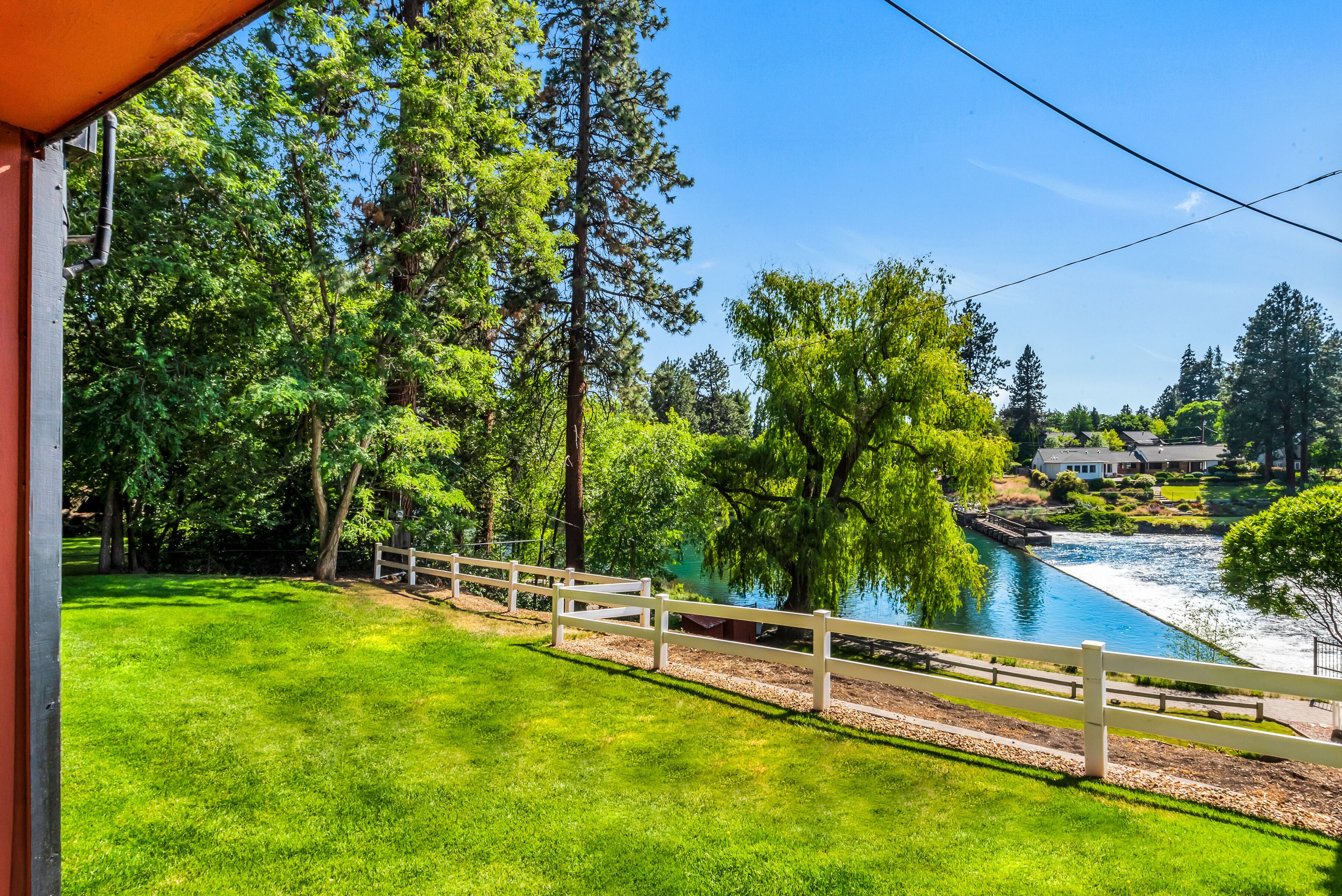 A riverside view with a wooden fence, lush greenery, and a house in the background.