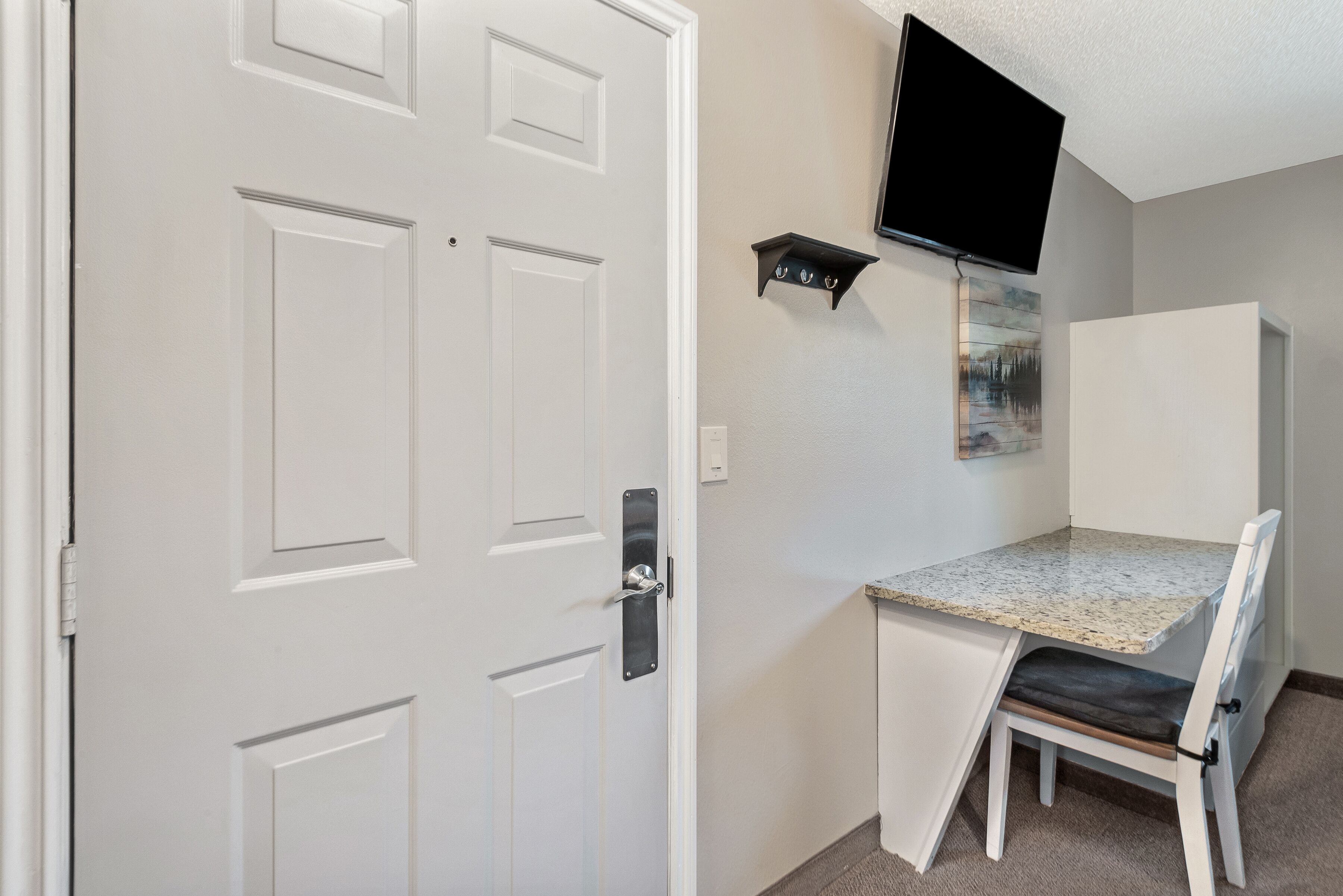 A small room with a white door, a television mounted on the wall, a small table with a chair, and a cabinet.