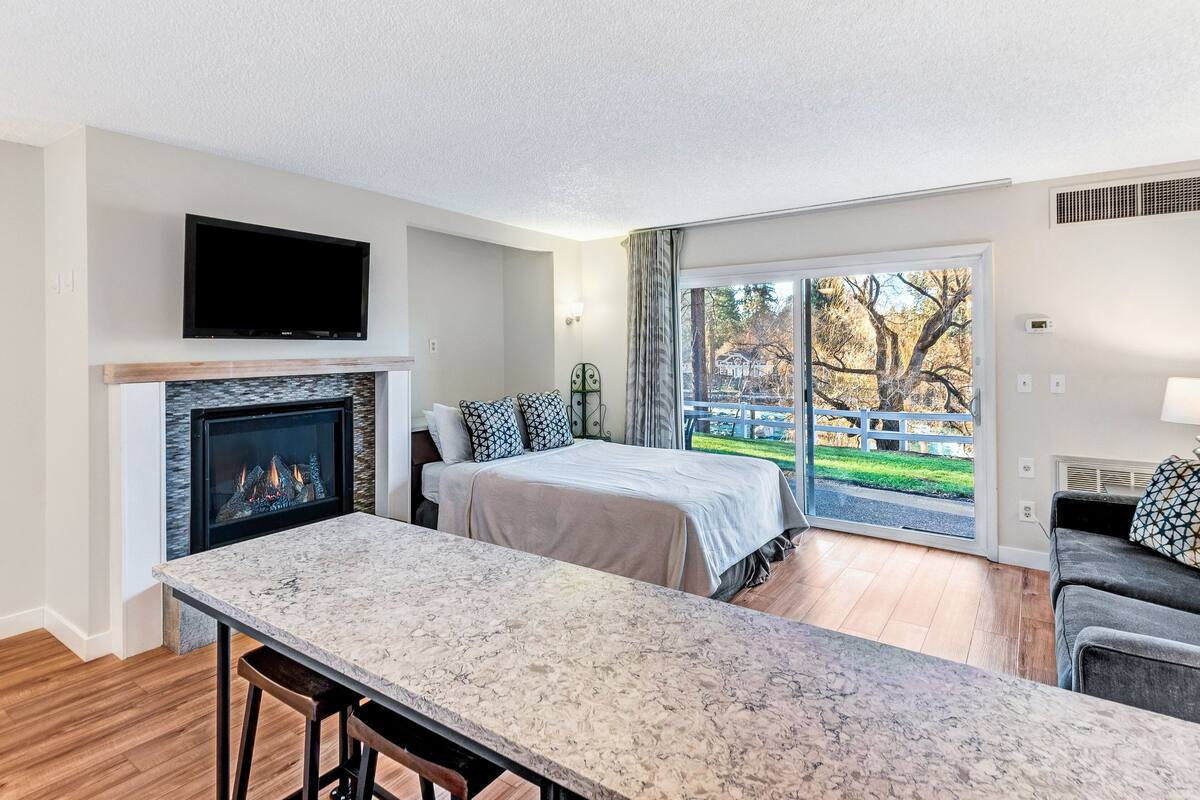 A modern hotel room with a fireplace, a flat-screen TV, a large bed, a dining area, and a view of a park.