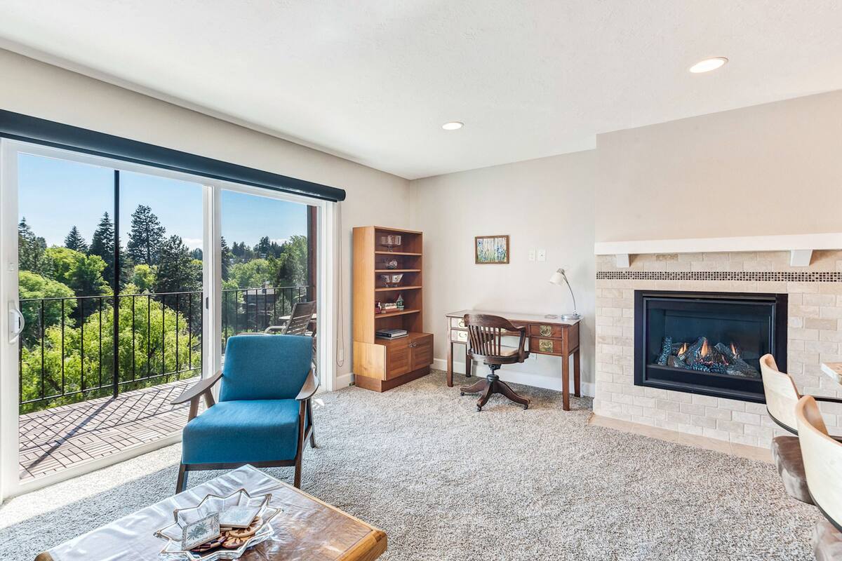 A spacious living room with a large window, a fireplace, a bookshelf, and a blue chair.