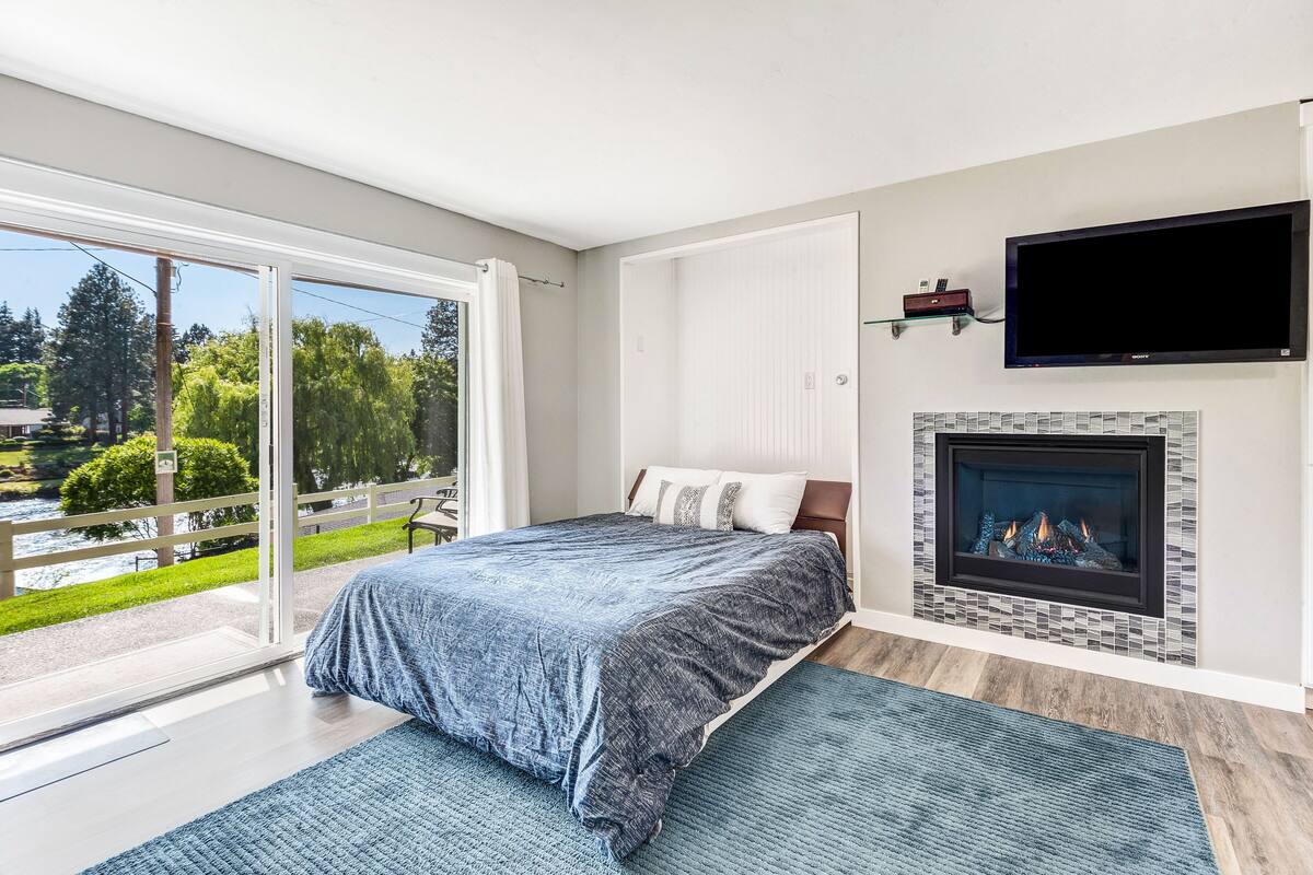 A bedroom with a large bed, a fireplace, a TV mounted on the wall, and a sliding glass door leading to a balcony with a view of greenery and a body of water.