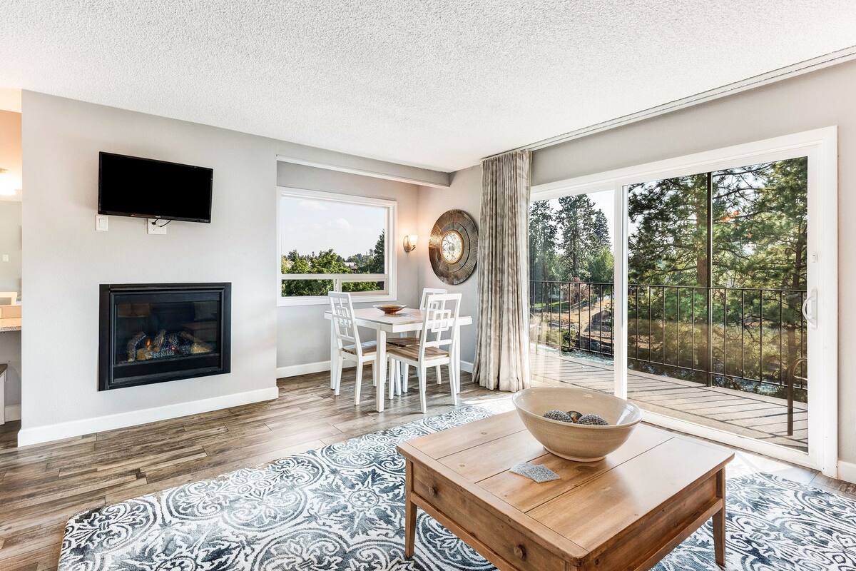 A modern living room with a fireplace, a dining area, and a large sliding glass door leading to a balcony with a view of trees and a lake.