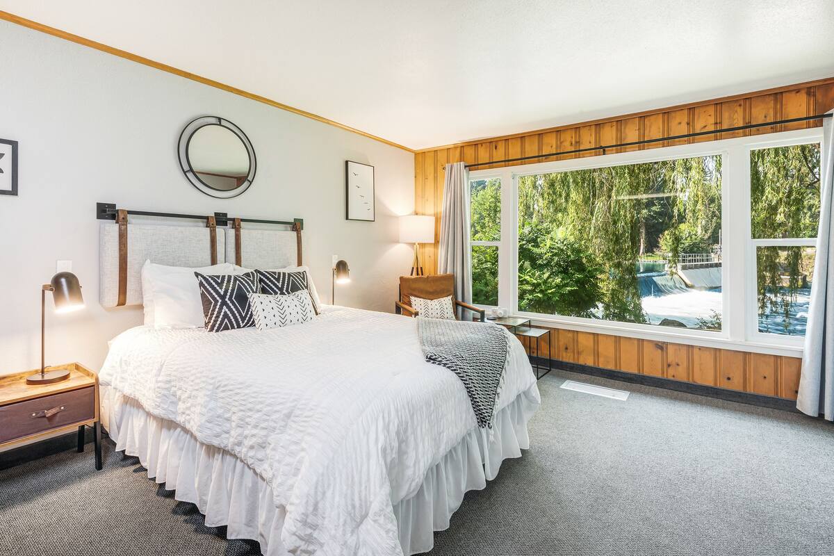 A bedroom with a large bed, a nightstand, a bedside table, a lamp, a window with a view of trees and a car outside.