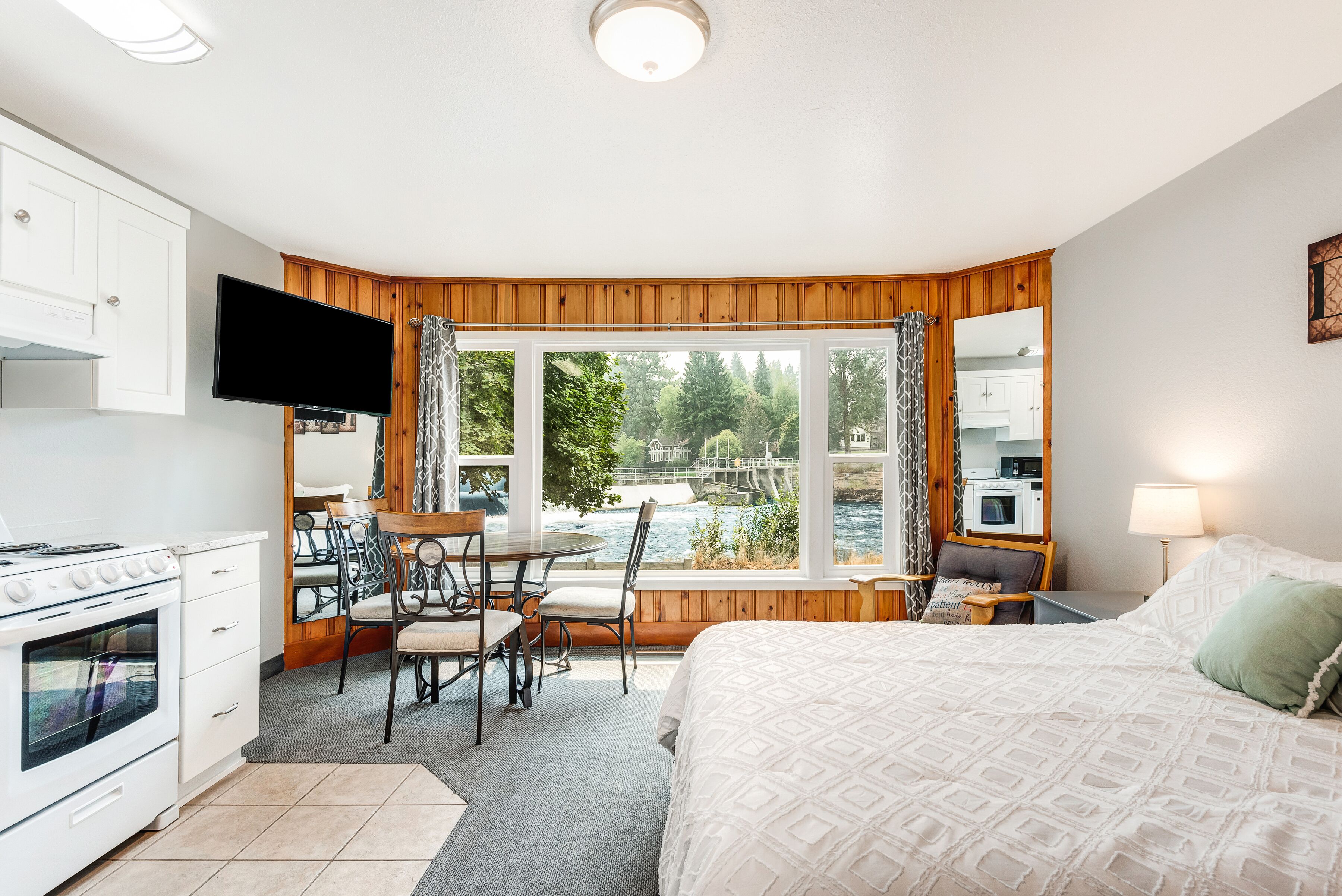 A bedroom with a bed, a TV, a dining area with chairs, a kitchen with an oven, and a large window with a view of a bridge and greenery.