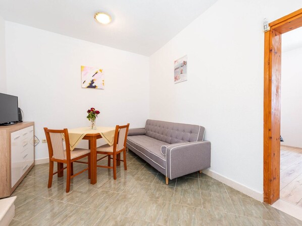 Une petite pièce bien éclairée, avec une table à manger, deux chaises, un canapé et un téléviseur posé sur un meuble en bois.