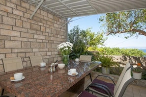 An outdoor dining area with a stone wall, a long table, and chairs. There are potted plants and a view of the sea. - Apartments Mira -one Bedroom Apartment With Terrace and Sea View (Supetar)