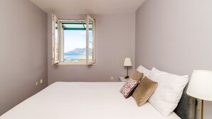 A bedroom with a large bed, a window with an open view of the sea, a bedside table with a lamp, and a nightstand. - Apartment Radovan - One-bedroom Apartment With Balcony and Sea View (Dubrovnik)