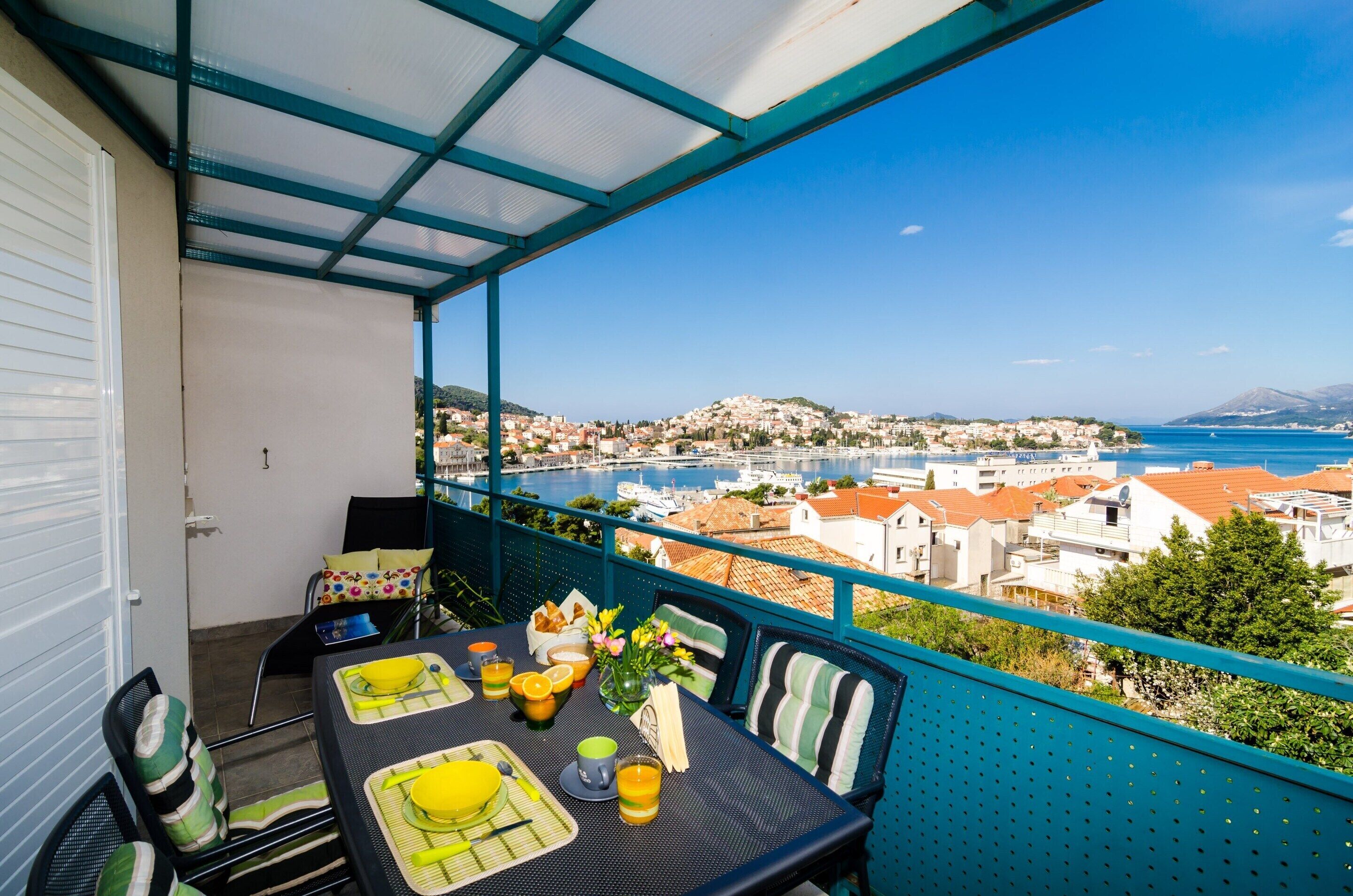 A balcony with a table set for breakfast, overlooking a coastal town and the sea.