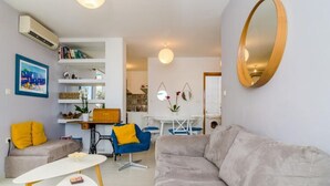 A modern living room with a grey sofa, a blue chair, and a white coffee table. There is a dining area with a white table and chairs, and a kitchen area with a stove and cabinets. - Apartment Radovan - One-bedroom Apartment With Balcony and Sea View (Dubrovnik)