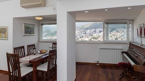 A dining area with a table set for four, a piano, and a view of a cityscape through large windows. - Apartment Super Marijo - Two-bedroom Apartment With Sea View (Dubrovnik)