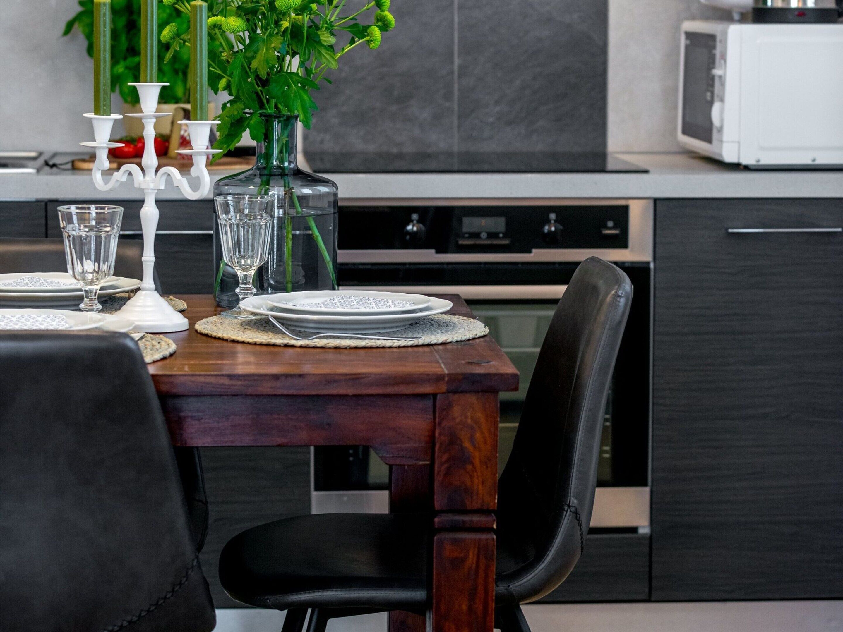 A modern kitchen with dark cabinetry, a wooden dining table set with plates, glasses, and a vase with green plants.