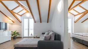 A modern loft bedroom with exposed wooden beams, a large window, a sofa bed, a TV, and a potted plant. - Apartments Terra - Comfort One Bedroom Apartment With Balcony and City View (Dubrovnik)