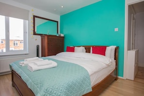 Une chambre avec un mur de couleur turquoise, un lit en bois, du linge de lit blanc et une fenêtre donnant sur des immeubles.