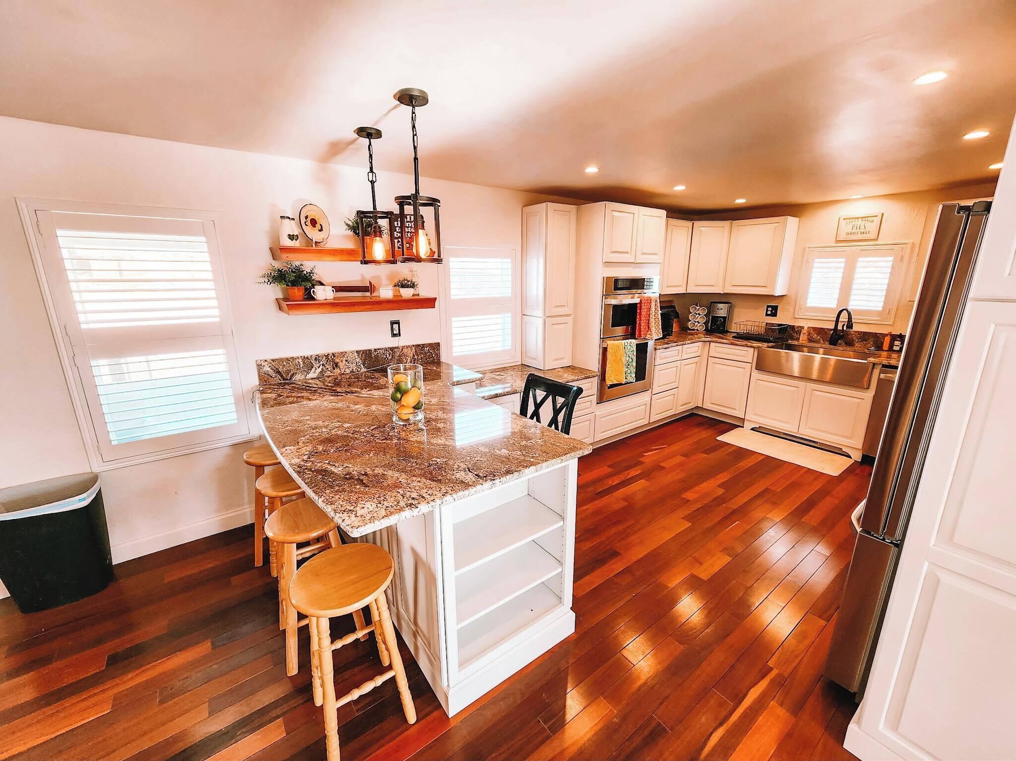 Una cocina con encimera de granito, taburetes de madera, armarios blancos, un lavabo de acero inoxidable y un refrigerador.