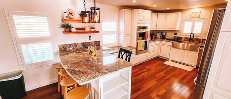 Una cocina con encimera de granito, taburetes de madera, armarios blancos, un lavabo de acero inoxidable y un refrigerador.