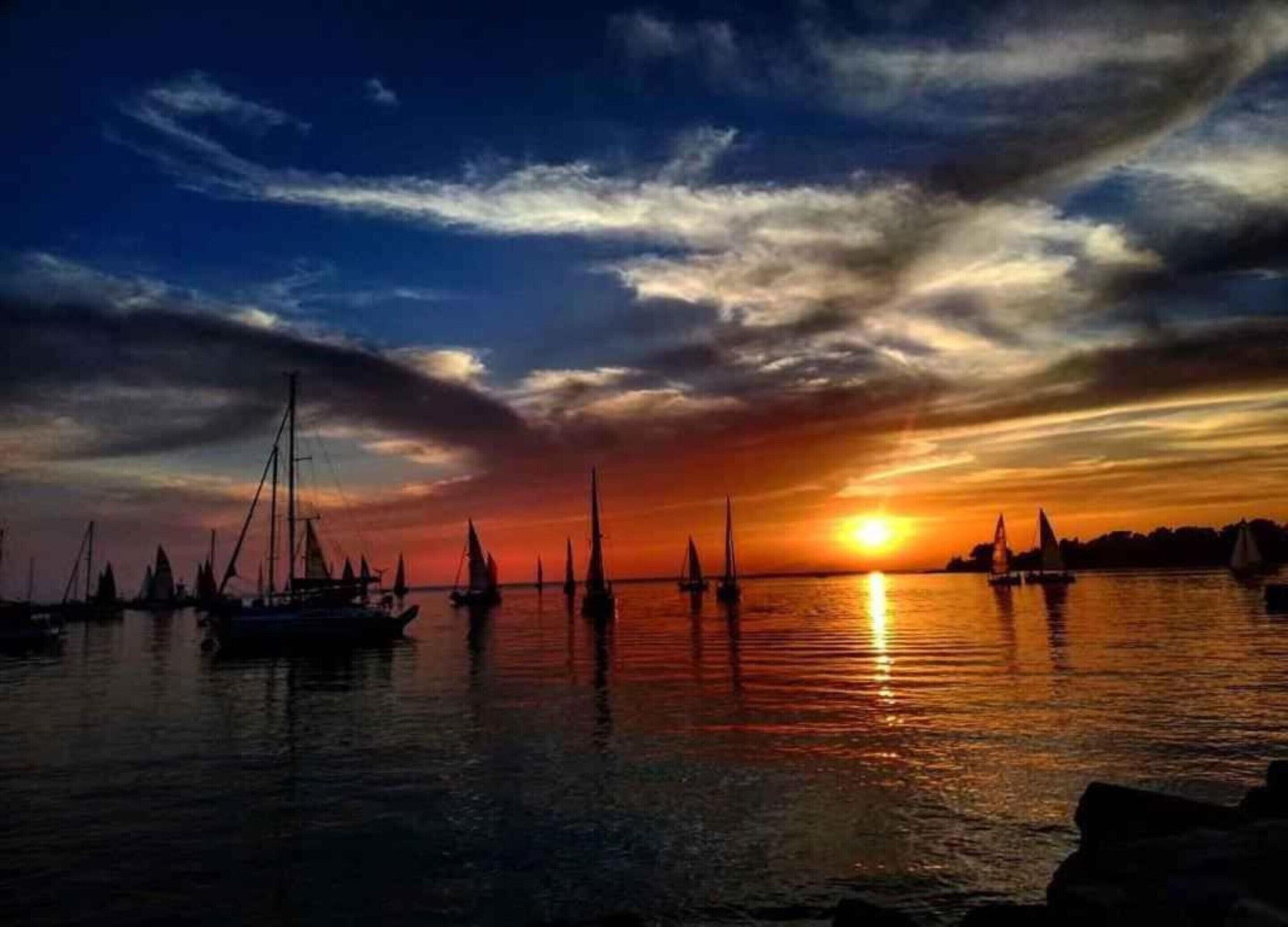 A sunset over a calm harbor with several sailboats anchored.