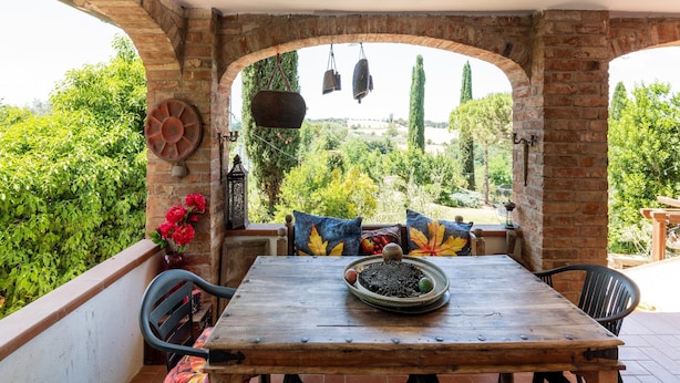 A wooden table with a bowl and a hanging light fixture in a covered outdoor area with brick arches and a view of greenery.