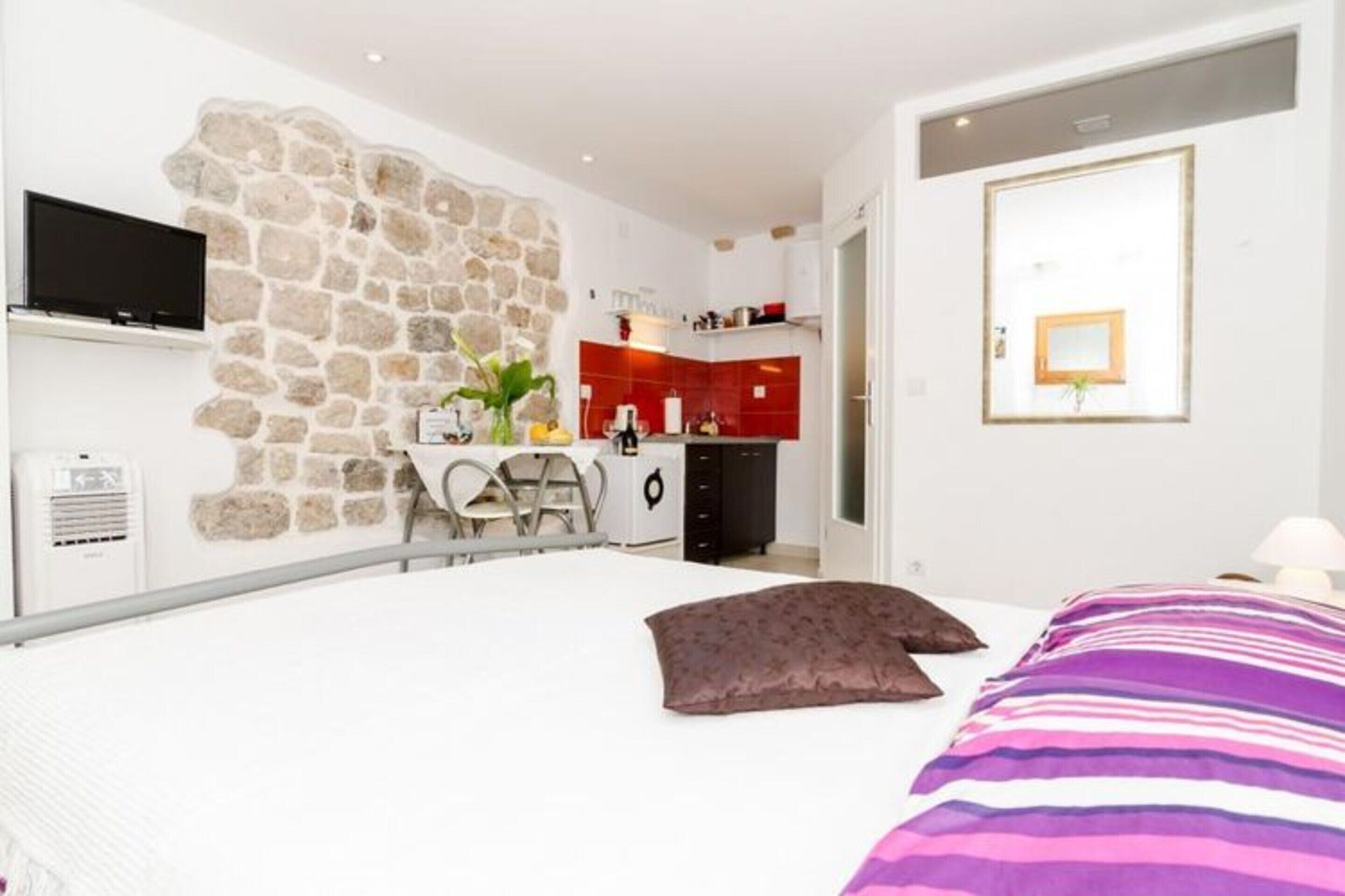 Une chambre avec un lit, un mur en pierre, une télévision, un bureau et, au fond, un coin cuisine.
