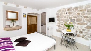 A bedroom with a bed, a small table with chairs, a TV, and a stone wall.