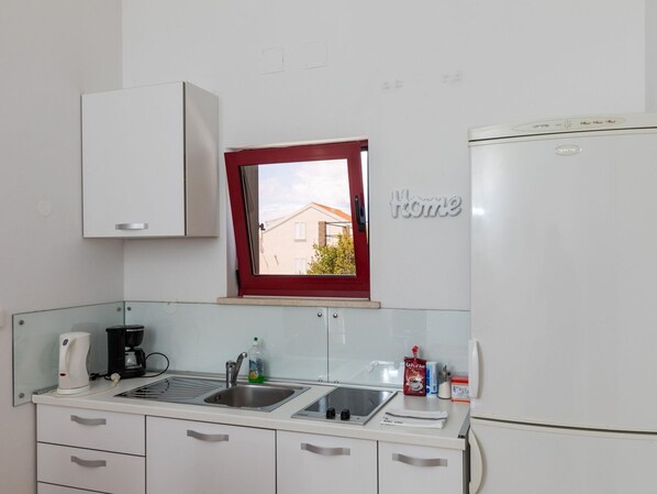 A compact kitchen with white cabinets, a built-in sink, a window with a red frame, and a refrigerator. - Apartments Franka - One Bedroom Apartment With Terrace and Garden View (Dubrovnik)